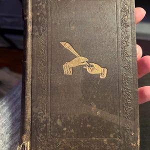 Op de afbeelding: Een vintage, bruin boek met gouden details. De omslag toont een gouden illustratie van twee handen, één met een penseel en de andere met een palet. De randen van het boek zijn versierd met een bloemmotief.