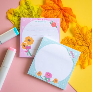 Puede incluir: Dos blocs de notas rosas y azules con diseños florales y el texto "Notas para un día florido" y "Notas para recordar relajarse". Los blocs de notas están sobre un fondo rosa y amarillo con hojas de otoño.
