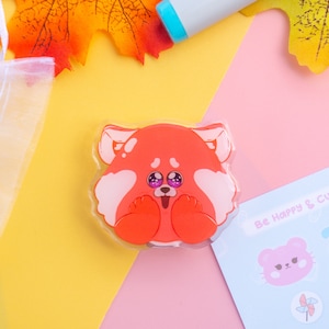 Op de afbeelding: Een transparante acrylbedel met een rood panda-personage met grote ogen en een roze neus. De bedel bevindt zich op een gele en roze achtergrond met een stickerblad waarop "Be Happy & Cutiey Stationery" staat.