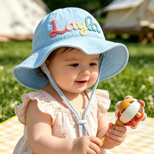 May include: A light blue baby sun hat with the name "Layla" embroidered in colorful thread. The hat has a wide brim and a chin strap. The baby is wearing a floral romper and holding a rattle.
