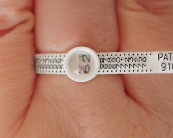 Pode incluir: Um medidor de tamanho de anel de plástico branco com uma lupa que mostra o tamanho "20". O medidor tem marcas pretas e o texto "PAT. PEND. 9101693".