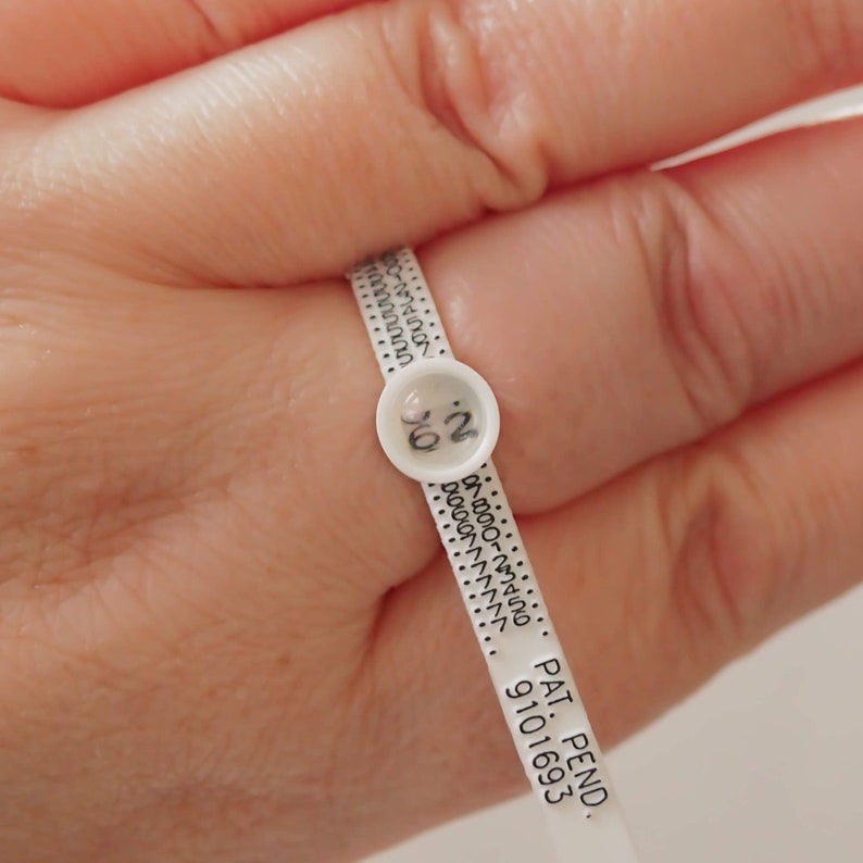 Puede incluir: Calibre de tama&ntilde;o de anillo de pl&aacute;stico blanco con marcas negras y una peque&ntilde;a etiqueta blanca con el n&uacute;mero "0.2" impreso en ella. El texto "PAT. PEND. 9101693" est&aacute; impreso en el anillo.
