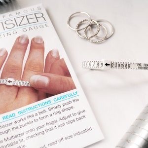 Puede incluir: Un medidor de talla de anillo de pl&aacute;stico blanco con una flecha negra y n&uacute;meros del 4 al 17. El medidor de talla de anillo est&aacute; etiquetado como "Multisizer" y "Ring Sizing Gauge". El texto "Lea atentamente las instrucciones" est&aacute; impreso en la tarjeta blanca debajo del medidor de talla de anillo.