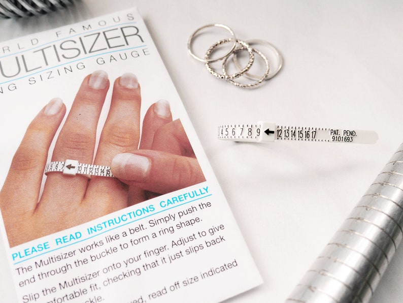 Puede incluir: Un medidor de talla de anillo de pl&aacute;stico blanco con una flecha negra y n&uacute;meros del 4 al 17. El medidor de talla de anillo est&aacute; etiquetado como "Multisizer" y "Ring Sizing Gauge". El texto "Lea atentamente las instrucciones" est&aacute; impreso en la tarjeta blanca debajo del medidor de talla de anillo.
