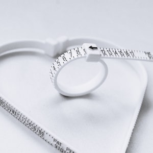 Puede incluir: Un medidor de talla de anillo de pl&aacute;stico blanco con los n&uacute;meros del 1 al 17 impresos en &eacute;l. El medidor tiene la forma de un anillo y se utiliza para medir el tama&ntilde;o de un dedo.