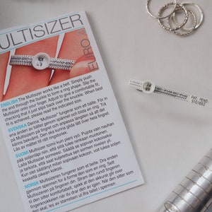 Puede incluir: Un ajustador de anillos de pl&aacute;stico blanco con marcas negras y el texto "Multisizer" y "Euro" impreso en &eacute;l. El ajustador est&aacute; dise&ntilde;ado para medir los tama&ntilde;os de los anillos y se muestra sobre una superficie blanca.