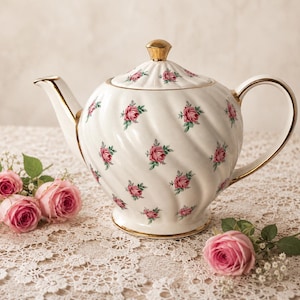 Puede incluir: Tetera de porcelana blanca con detalles dorados y tapa, adornada con motivos de rosas rosas. La tetera está sobre un mantel de encaje, con rosas rosas y follaje cerca. La tetera mide aproximadamente 20 cm de altura.