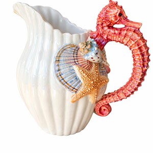 May include: White ceramic pitcher with a seahorse handle in shades of red and orange. The pitcher is decorated with seashells and a starfish. The pitcher has a scalloped rim and a pearlescent finish, perfect for coastal decor.