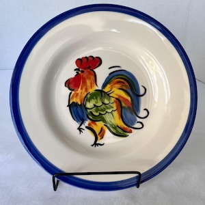 May include: A white ceramic plate with a hand-painted rooster design. The rooster features red, yellow, green, and blue colors. The plate has a blue border and is displayed on a black metal stand.
