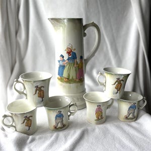 May include: A ceramic pitcher and five matching mugs featuring illustrations of figures in traditional clothing. The pitcher displays a group of three people, while each mug shows a single figure in colorful attire. The set has a cream base color.