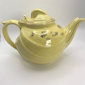 Puede incluir: Una tetera de cerámica de color amarillo pálido con tapa y asa en espiral. La tetera tiene adornos dorados y diseños de hojas doradas. El pico es curvo y el cuerpo tiene un diseño acanalado.
