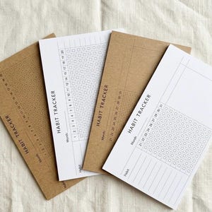 May include: Four habit tracker notepads. Two are kraft paper, and two are white. Each notepad has a grid for tracking habits, with the words "Habit Tracker" printed vertically. The kraft paper notepads have a grid for 20 days, and the white ones have a grid for 31 days.