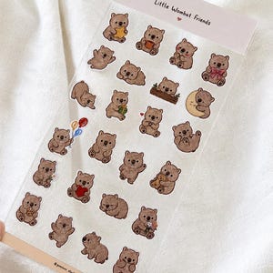 Op de afbeelding: Een transparant stickervel met de titel Little Wombat Friends met talloze kleine, bruine, geïllustreerde wombats in verschillende houdingen, waaronder met sterren, ballonnen, harten, cadeaus en bloemen, met de tekst gemtuni studio onderaan.