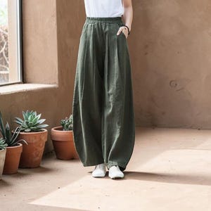 May include: Olive green wide-leg pants with a relaxed fit. The pants have a high waist and pockets. The model is wearing white sneakers and a white top. The pants are made of a soft, flowing fabric.