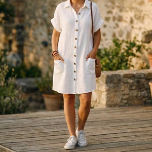 May include: A white, short-sleeved dress with a button-down front and two front pockets. The dress is paired with white sneakers. A brown woven bag hangs from a shoulder strap. The background features a stone wall and greenery.