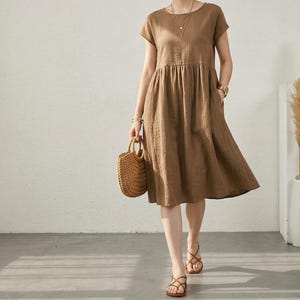 Pode incluir: Um vestido de linho castanho com mangas curtas e cintura franzida. O vestido chega ao joelho e é usado com sandálias de couro castanho e uma bolsa de mão de palha tecida. A pessoa usa joias douradas.