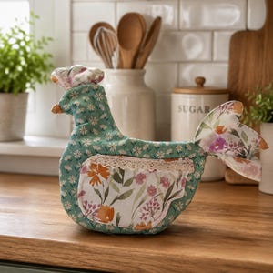 May include: A fabric chicken-shaped tea cozy featuring a teal background with a small white floral pattern. It includes a side pocket with a larger floral print in orange, pink, and green, accented by a cream-colored lace trim along the top edge.
