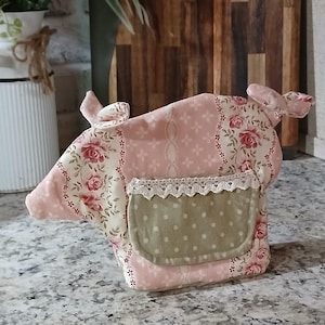 May include: A fabric pig-shaped ornament featuring a patchwork design of pink floral and geometric patterns. It includes a sage green polka-dot pocket on the side, accented with a delicate white lace trim along the top edge of the pocket.