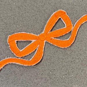 Puede incluir: Un parche bordado con forma de lazo naranja con ribete blanco, sobre un fondo gris moteado. El parche es un elemento decorativo, probablemente para ropa o manualidades.