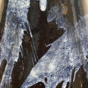 May include: Close-up of a ceramic vase with a dark brown base and a glossy, dark blue glaze. The glaze features a unique, textured pattern of white and blue drips and splatters, creating an abstract design.