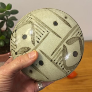 Puede incluir: Un recipiente cerámico redondo con un fondo verde claro y un patrón geométrico en tonos grises y negros. El recipiente tiene tapa y está sostenido por una mano. El diseño incluye líneas, puntos y formas. Probablemente sea para almacenamiento o exhibición.