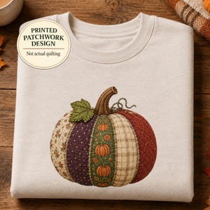 May include: A beige crewneck sweatshirt featuring a central graphic of a pumpkin with a printed patchwork design. The pumpkin is composed of various fabric patterns including floral, polka dot, plaid, and quilted textures. Text reads: PRINTED PATCHWORK DESIGN Not actual quilting.