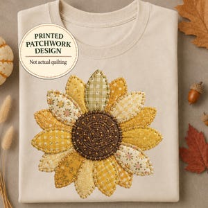 Puede incluir: Una camiseta beige con un gráfico de girasol grande con un diseño de patchwork impreso. El girasol tiene un centro marrón oscuro y pétalos con varios patrones amarillos y verdes. Una etiqueta circular dice: PRINTED PATCHWORK DESIGN Not actual quilting.