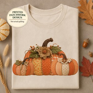 Puede incluir: Una camiseta beige de cuello redondo con un gráfico de tres calabazas con un diseño de patchwork impreso. Las calabazas están decoradas con patrones naranjas y amarillos, flores de tela y bayas. El texto dice: PRINTED PATCHWORK DESIGN Not actual quilting.