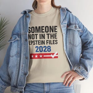 Może przedstawiać: Beżowa koszulka z czarnym napisem "SOMEONE NOT IN THE EPSTEIN FILES 2028". Poniżej czerwono-biało-niebieska grafika z gwiazdami i znakiem wyboru. Osoba ma na sobie niebieską kurtkę dżinsową.