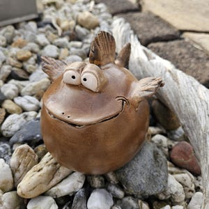 Könnte beinhalten: Eine skurrile, braune Keramikfischskulptur mit großen, ausdrucksstarken Augen und einem lächelnden Mund. Der Fisch hat Flossen und ist zwischen kleinen Steinen und Kieselsteinen platziert, was auf eine Gartendekoration im Freien hindeutet.
