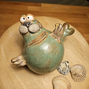 Könnte beinhalten: Eine skurrile Keramik-Robbenfigur in Grün- und Brauntönen, mit großen, ausdrucksstarken Augen und flossenartigen Armen. Die Robbe ist auf einer Holzoberfläche platziert, zusammen mit drei Muscheln.