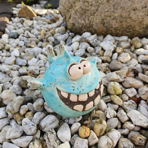 Może przedstawiać: Okrągła, jasnoniebieska ceramiczna figurka ryby z nakrapianym wykończeniem, posiadająca dwoje dużych, wypukłych oczu i szeroki, wyszczerzony uśmiech. Ryba ma małe płetwy po bokach i na grzbiecie oraz ciemnobrązowe szkliwo podkreślające usta.