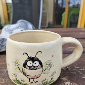 Könnte beinhalten: Ein cremefarbener Keramikbecher mit einer handgemalten Illustration einer runden, flauschigen Biene mit großen Augen, umgeben von weißen Gänseblümchen und grünem Gras. Die Biene hat kleine Flügel und ein winziges rotes Herz ist auf den Becher gemalt.