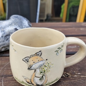 Könnte beinhalten: Ein cremefarbener Keramikbecher mit einer handgemalten Illustration eines kleinen orange-weißen Fuchses, der einen Strauß gelber Blumen hält. Der Fuchs sitzt auf einem grünen Grasbüschel, ergänzt durch kleine Blumenmotive an den Seiten.