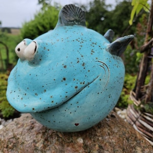 Pode incluir: Uma figura de peixe em cerâmica azul-clara, de forma redonda, com pintas castanhas escuras. O peixe apresenta dois olhos brancos proeminentes e salientes, um sorriso largo esculpido e barbatanas cinzentas escuras no topo e na lateral.
