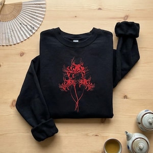 May include: Black sweatshirt with a red floral design. The crew neck sweatshirt has long sleeves with cuffed wrists. The design is a stylized illustration of red spider lilies. The sweatshirt is displayed on a light brown wooden surface.