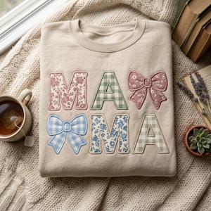 Op de afbeelding: Beige sweatshirt met het woord "MAMA" in bloemen- en ruitpatronen en strikaccenten. De letters zijn roze, groen, blauw en beige. De sweatshirt ligt op een crèmekleurige gebreide deken.