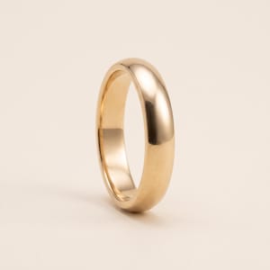 Può includere: Un anello in oro lucido con una superficie esterna liscia e arrotondata, caratterizzato da un design a fascia semplice e solido. Il metallo presenta una finitura calda e riflettente che cattura la luce, creando un aspetto uniforme e pulito.