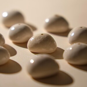 May include: A collection of off-white, rounded ceramic beads with a glossy finish. Each bead has a small hole at the top. The beads are arranged on a light beige surface, casting soft shadows.