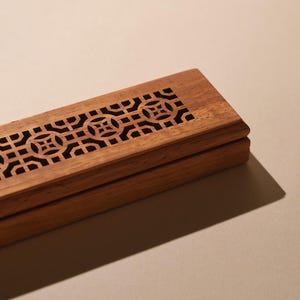 May include: A rectangular wooden box with a decorative geometric pattern carved into the lid. The box is made of light brown wood and has a dark interior. The design is a series of interlocking shapes.