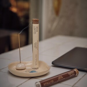 May include: A lit incense stick with smoke rising, next to a tube labeled "QUIET RISE" and "natural agarwood". A second tube reads "WARM DUSK". The incense sits on a small, off-white ceramic dish. A laptop is in the background.