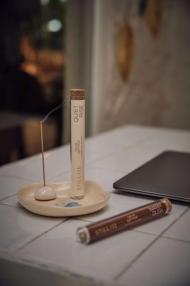 May include: A lit incense stick with smoke rising, next to a tube labeled "QUIET RISE" and "natural agarwood". A second tube reads "WARM DUSK". The incense sits on a small, off-white ceramic dish. A laptop is in the background.