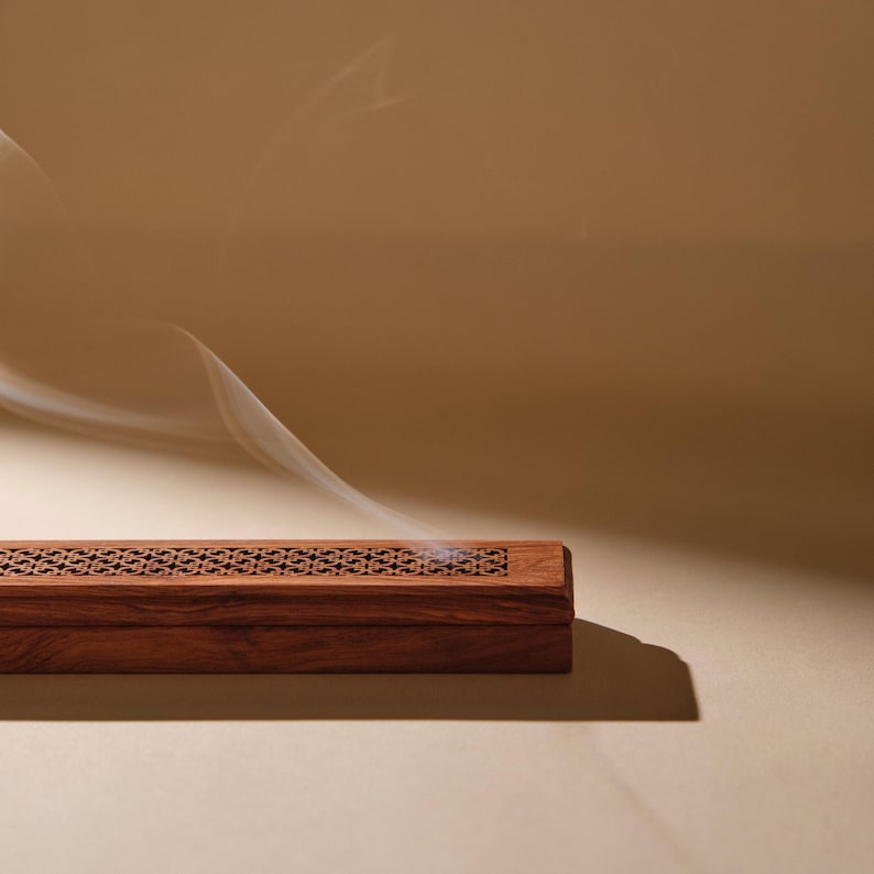 May include: A rectangular, wooden incense burner with intricate carved patterns on the top. The dark brown box emits a wisp of white smoke against a warm, neutral background, suggesting a calming atmosphere.