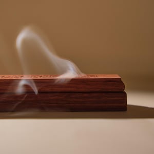 May include: A rectangular, dark brown wooden incense burner with carved details. Wisps of white smoke rise from the top, suggesting burning incense. The burner is set against a neutral background, highlighting its rich color and design.