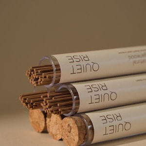 May include: Three tubes of incense sticks with cork stoppers. The tubes are labeled "QUIET RISE" in a stacked format. The incense sticks are brown, and the tubes are clear. The background is a solid, neutral color.