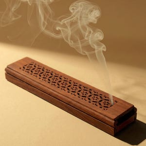 May include: A rectangular, wooden incense holder with a decorative, geometric cutout pattern on the lid. Wisps of white smoke rise from the holder, suggesting burning incense. The holder is a rich, dark brown color.