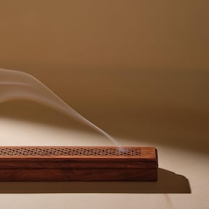 May include: A rectangular, wooden incense burner with intricate carved patterns along the top edge. Wisps of white smoke emanate from the burner, creating a sense of tranquility. The wood has a rich, dark brown color, and the background is a warm, neutral tone.