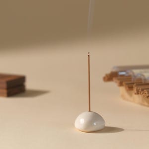 May include: A lit incense stick in a white ceramic holder, with smoke rising. A wooden block and a package of incense sticks are in the background. The scene is set on a neutral-toned surface.