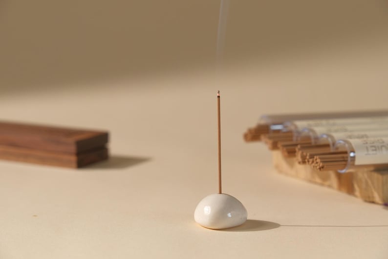 May include: A lit incense stick in a white ceramic holder, with smoke rising. A wooden block and a package of incense sticks are in the background. The scene is set on a neutral-toned surface.