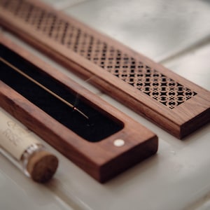 May include: A wooden incense holder with a decorative, geometric pattern on the lid. The holder is open, revealing a lit incense stick. A glass tube with text is in the foreground, and the holder is on a tiled surface.